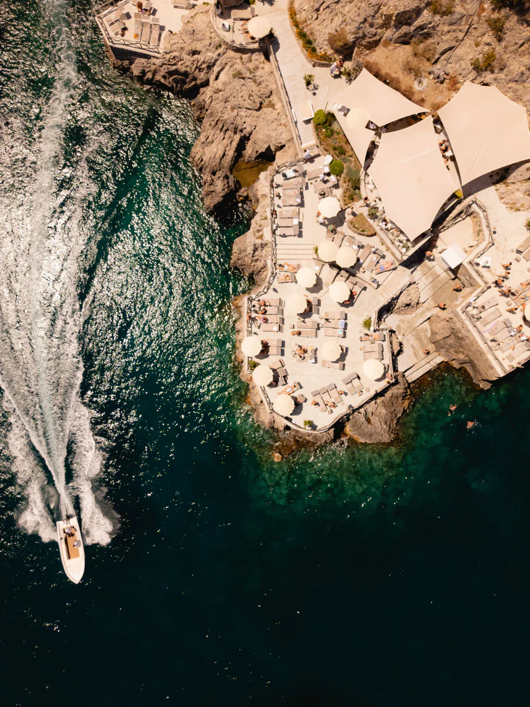 Aerial view of Conca del Sogno restaurant on the Amalfi Coast with sunbathers and a boat navigating clear waters.